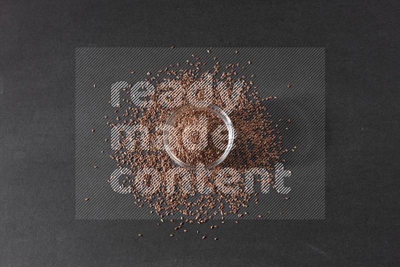 A glass bowl full of flaxseeds surrounded by the seeds on a black flooring