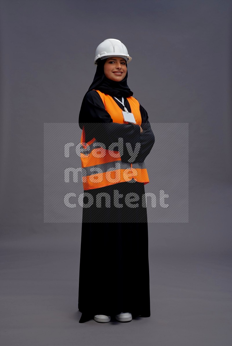 Saudi woman wearing Abaya with engineer vest with neck strap employee badge standing with crossed arms on gray background