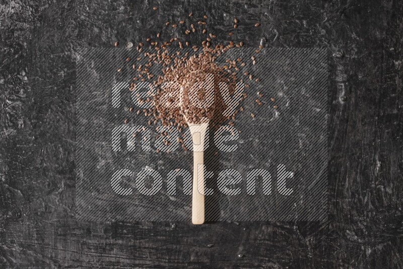 A wooden spoon full of flaxseeds and surrounded by seeds on a textured black flooring