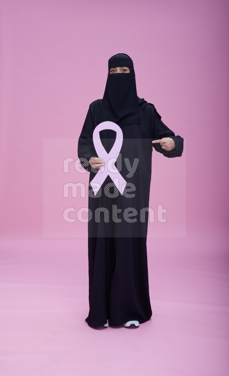 Saudi woman wearing abaya and niqab standing holding awareness ribbon on pink background