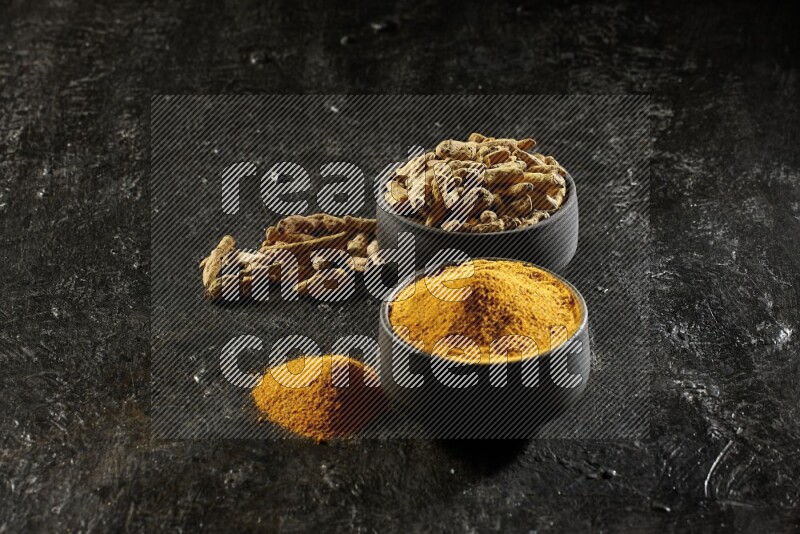 2 pottery black bowls, one full of turmeric powder and the other full of dried turmeric whole fingers on a textured black flooring