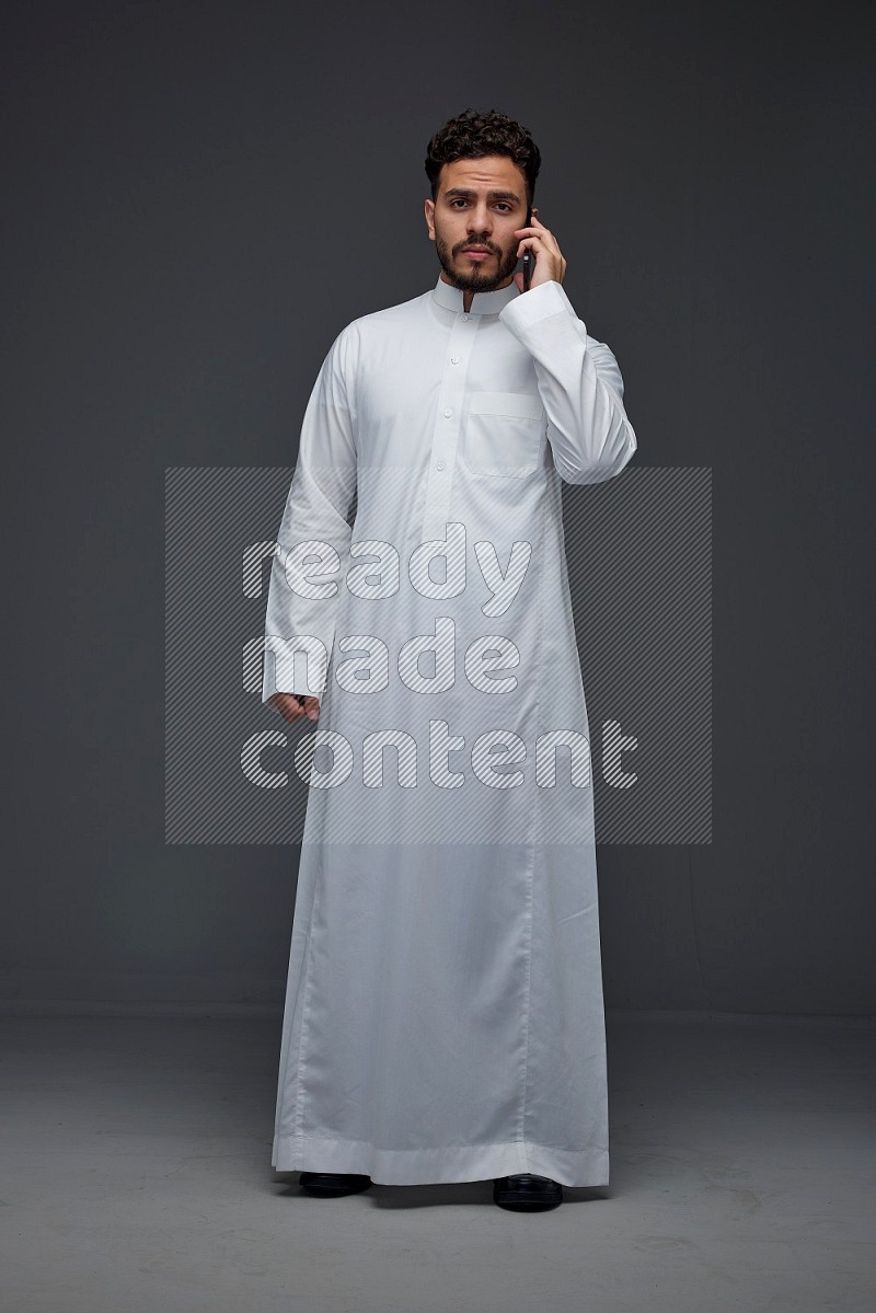 A Saudi man wearing Thobe and talking in the phone while standing and making different hand gestures eye level on a gray background
