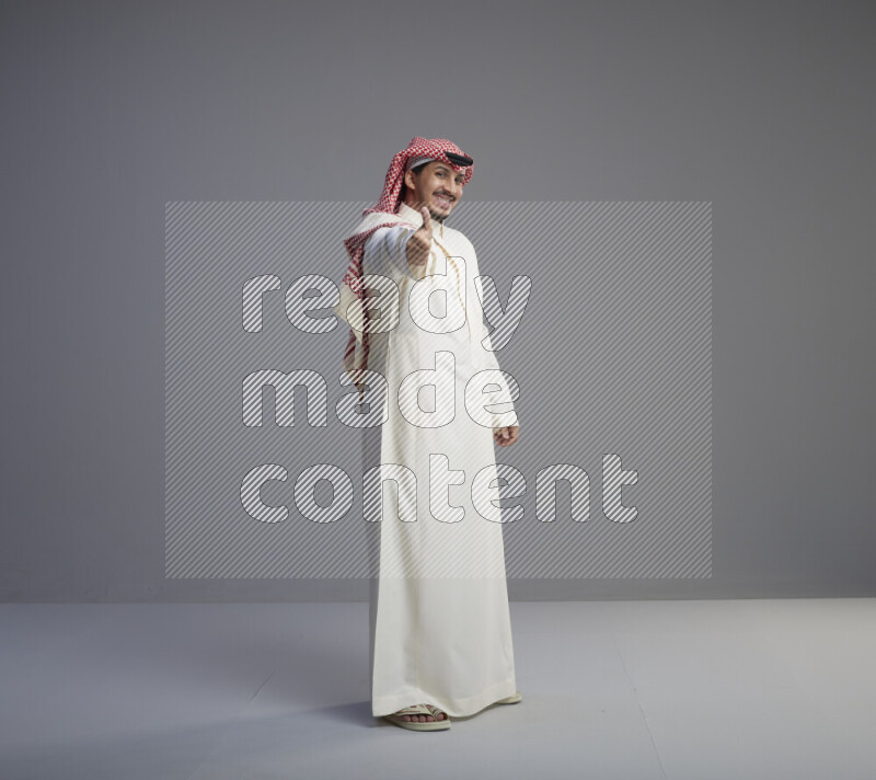 A Saudi man standing wearing thob and red shomag interacting with the camera on gray background