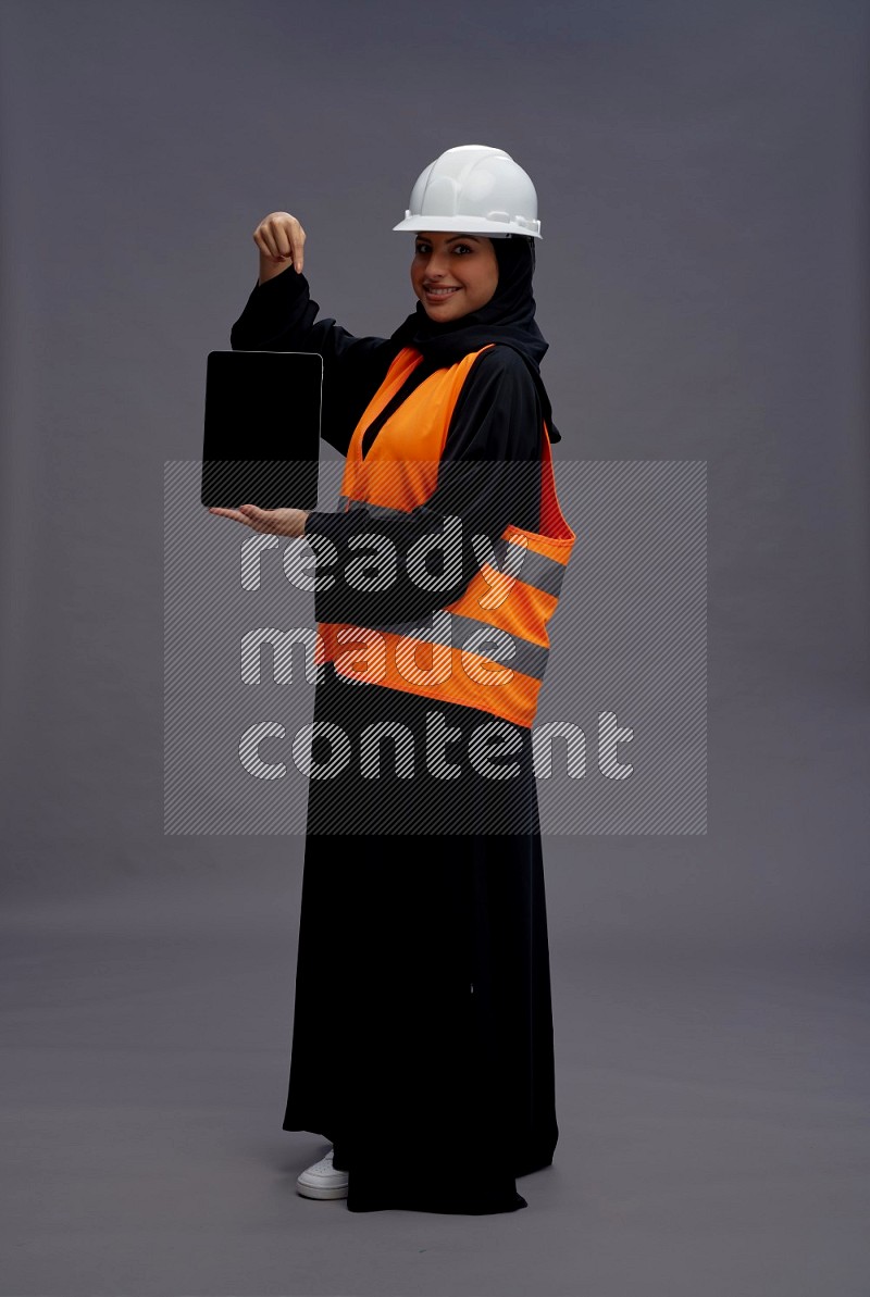 Saudi woman wearing Abaya with engineer vest standing showing tablet to camera on gray background