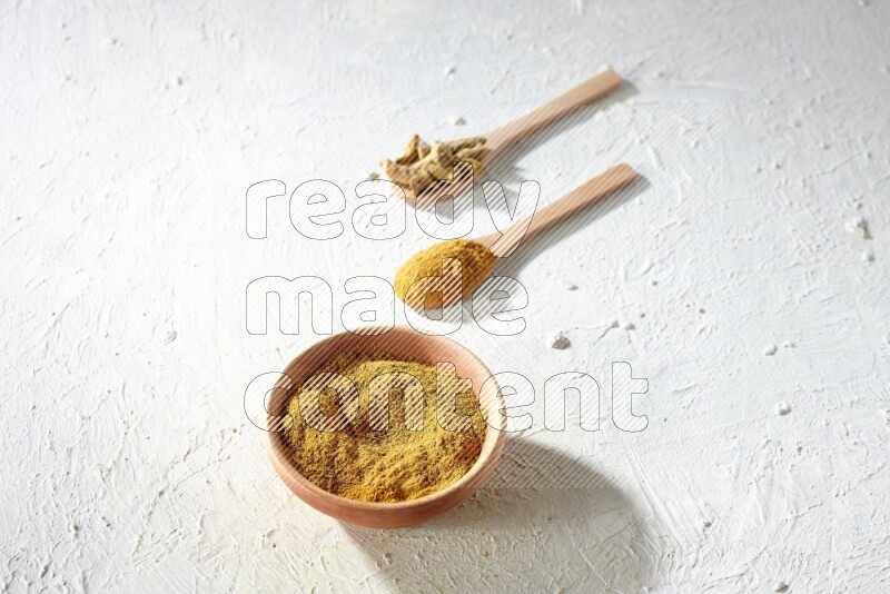 A wooden bowl full of turmeric powder and 2 wooden spoons full of dried turmeric whole finger and turmeric powder on textured white flooring