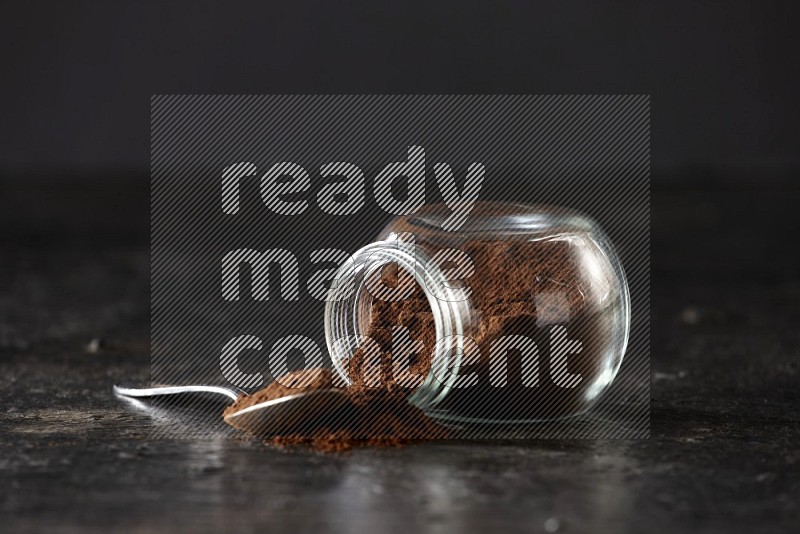 وعاء زجاجي للبهارات مقلوب وملعقة معدنية ممتلئان ببودرة القرنفل على أرضية سوداء