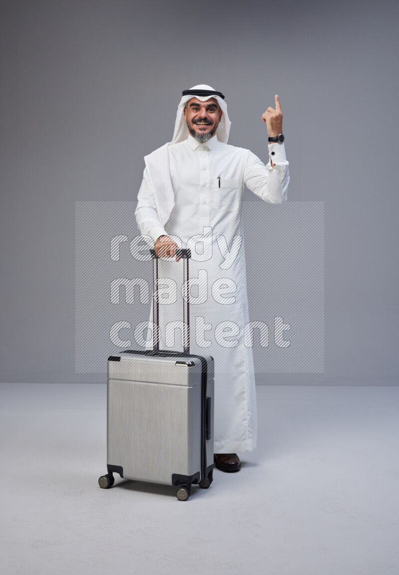 Saudi man wearing Thob and white Shomag standing holding Travel bag on Gray background