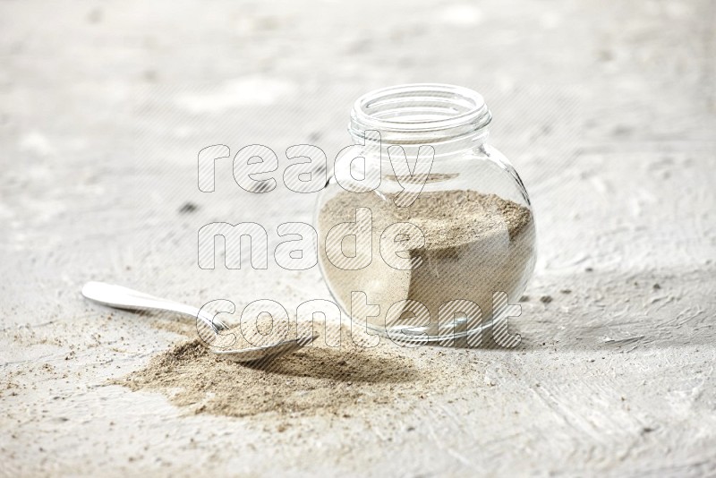 وعاء زجاجي عشبي ممتلئ ببودرة الفلفل الأبيض وملعقة معدنية ممتلئة بالبودرة على أرضية بيضاء