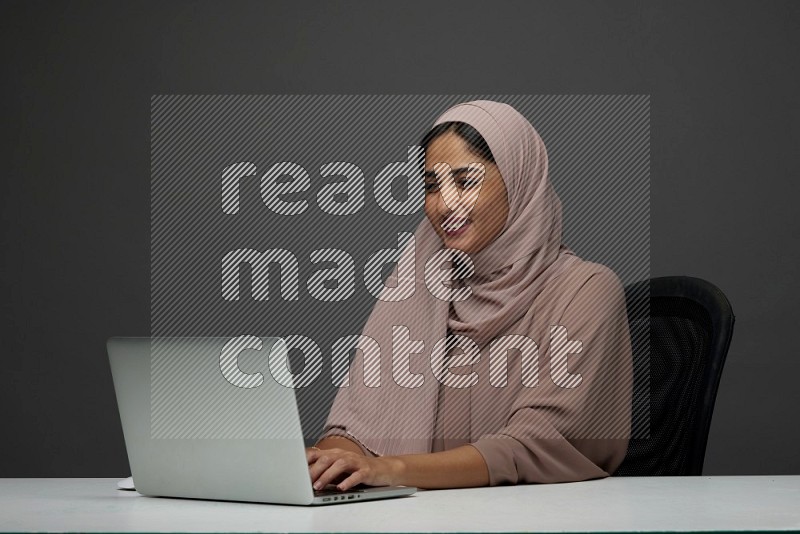 A Saudi woman Setting on her desk Typing on her laptop on a Gray Background wearing Brown Abaya with Hijab