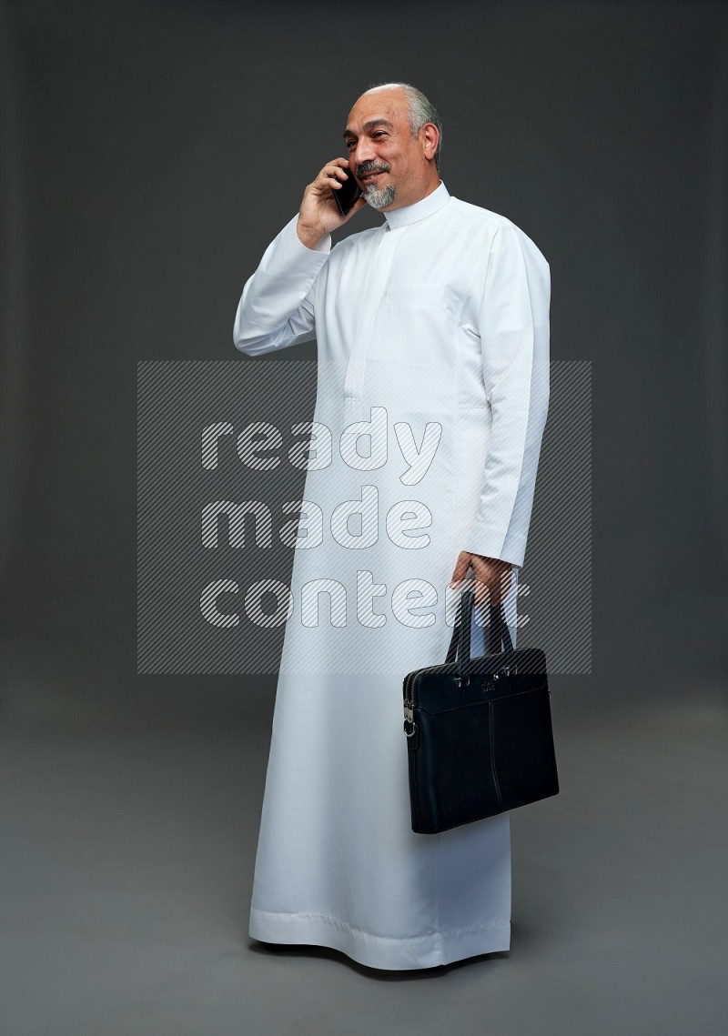 Saudi man without shomag Standing holding bag and talking on phone on gray background
