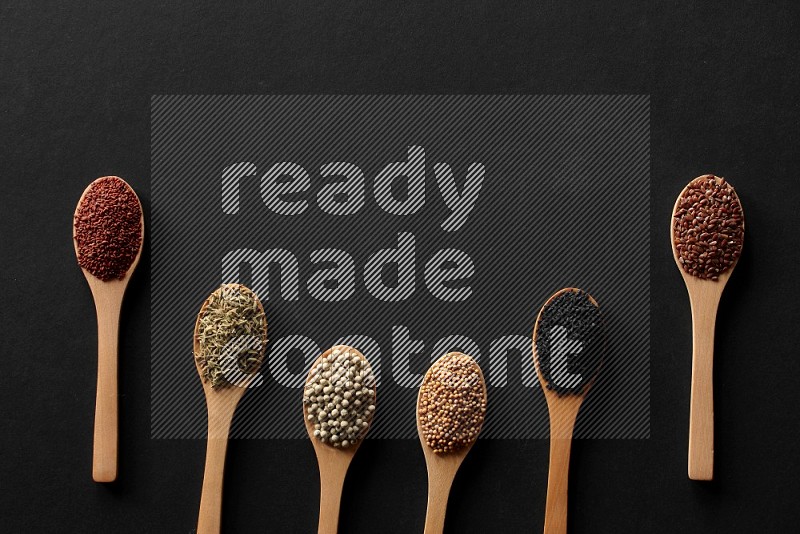 wooden spoons filled with white peppers, mustard seeds, black seeds, garden cress, cumin and flax on black flooring