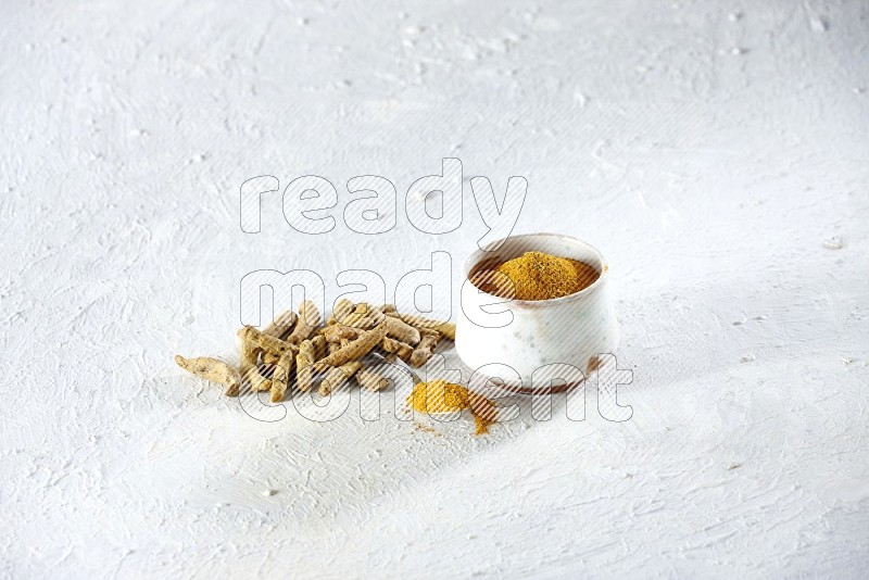 A beige pottery bowl and metal spoon full of turmeric powder and dried turmeric fingers next of them on textured white flooring