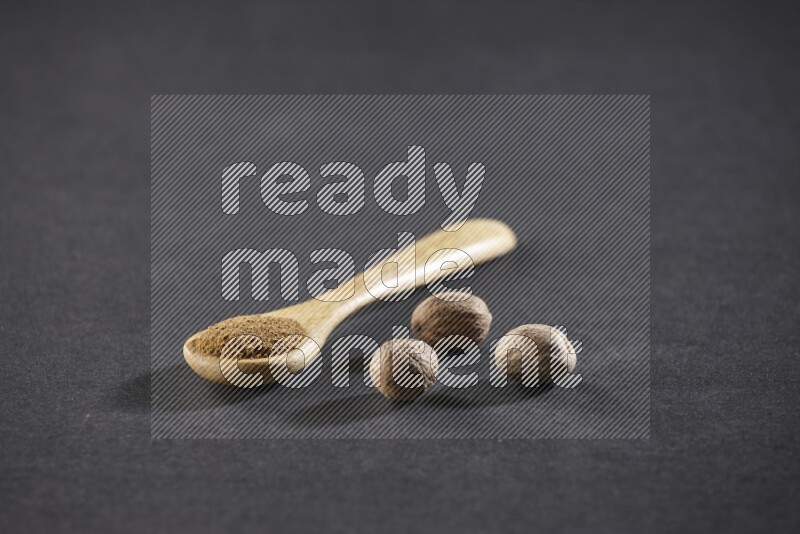 A wooden spoon full of nutmeg powder with whole nutmeg seeds beside it on a black flooring