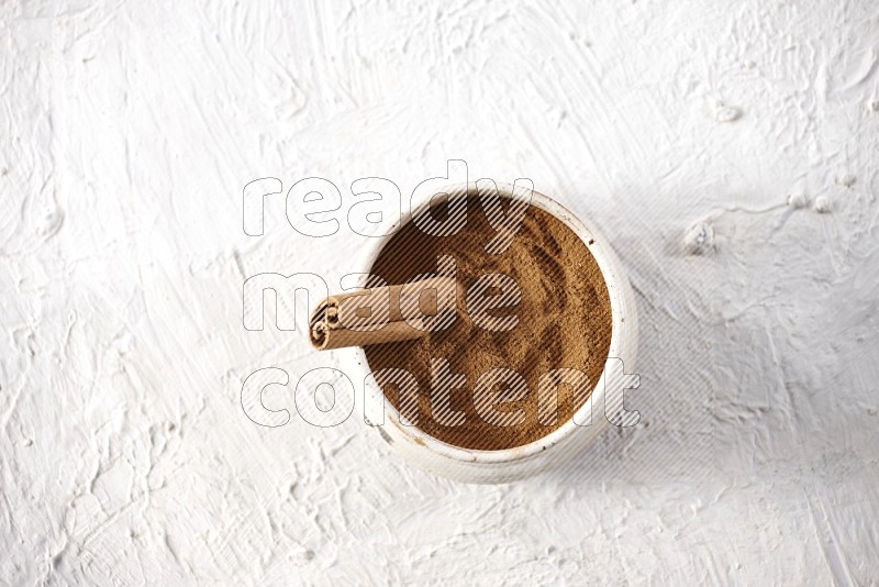 Ceramic beige bowl full of cinnamon powder with a cinnamon stick on a textured white background