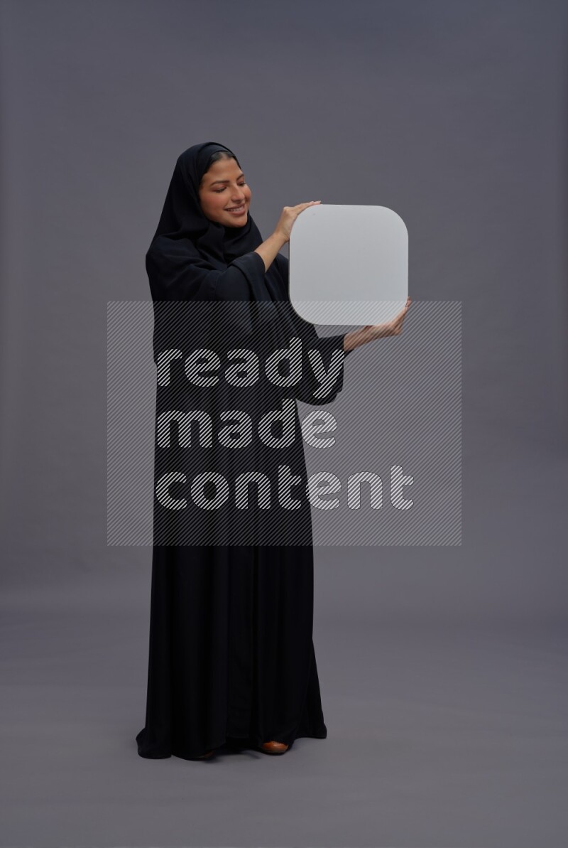 Saudi woman wearing Abaya standing holding social media sign on gray background