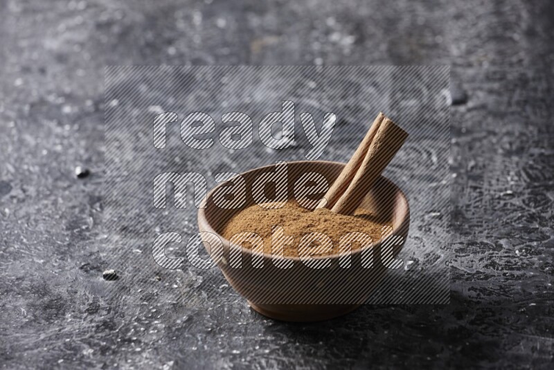 Wooden bowl full of cinnamon powder and a cinnamon stick on a textured black background