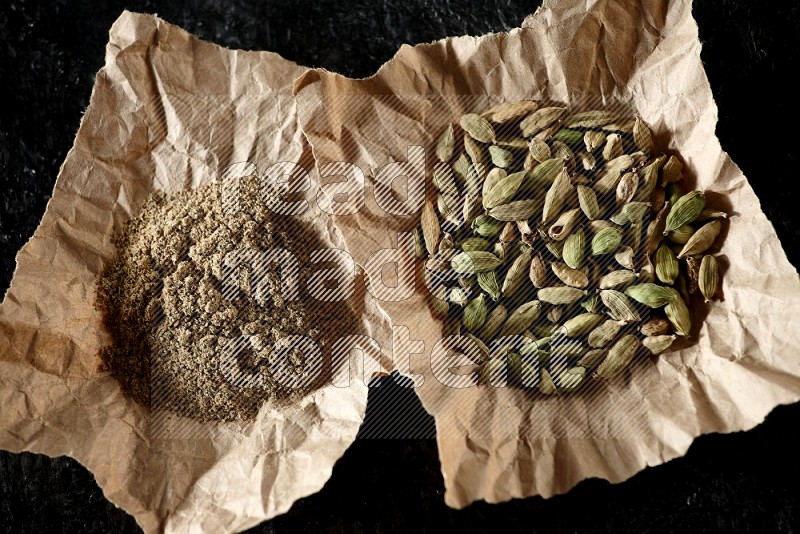 Cardamom seeds and cardamom powder in 2 crumpled pieces of paper on textured black flooring