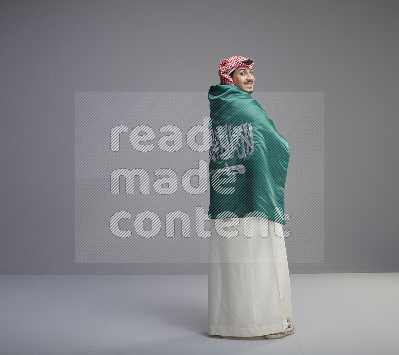 رجل سعودي يرتدي ثوب ابيض وشماغ احمر يحمل العلم السعودي على كتفيه