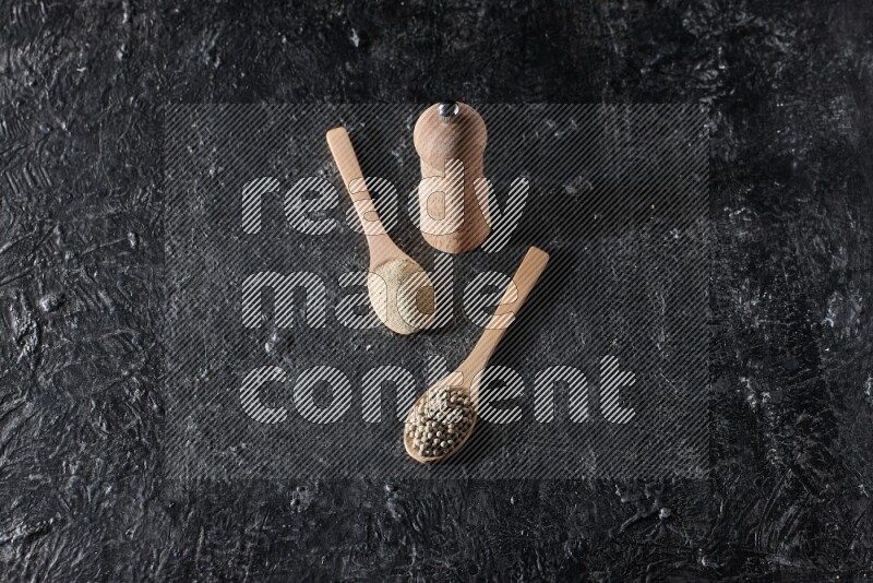 2 wooden spoons one full of white pepper powder and the other with pepper beads and a wooden pepper mill on textured black flooring