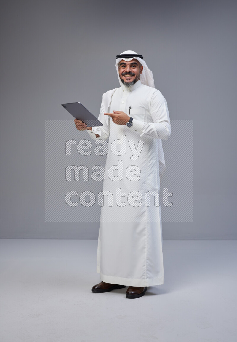 Saudi man Wearing Thob and white Shomag standing working on tablet on Gray background