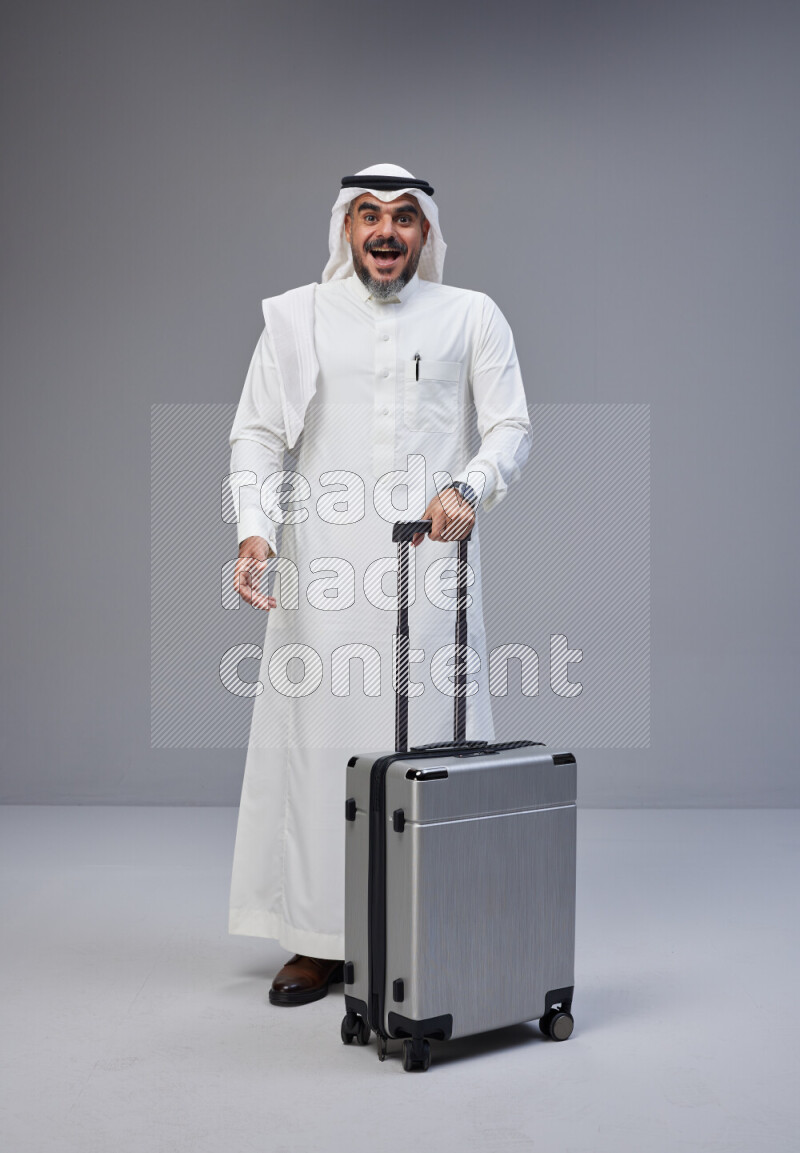 Saudi man wearing Thob and white Shomag standing holding Travel bag on Gray background