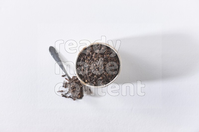 وعاء بيج سيراميك مملوء بالقرنفل وبجانبه ملعقة معدنية على أرضية بيضاء