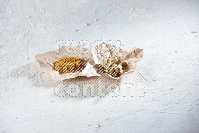 Turmeric powder and dried turmeric whole fingers in 2 crumpled pieces of paper on textured white flooring
