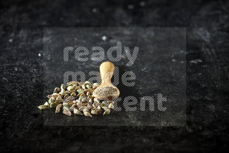 A wooden spoon full of cardamom powder and cardamom seeds next of it on textured black flooring