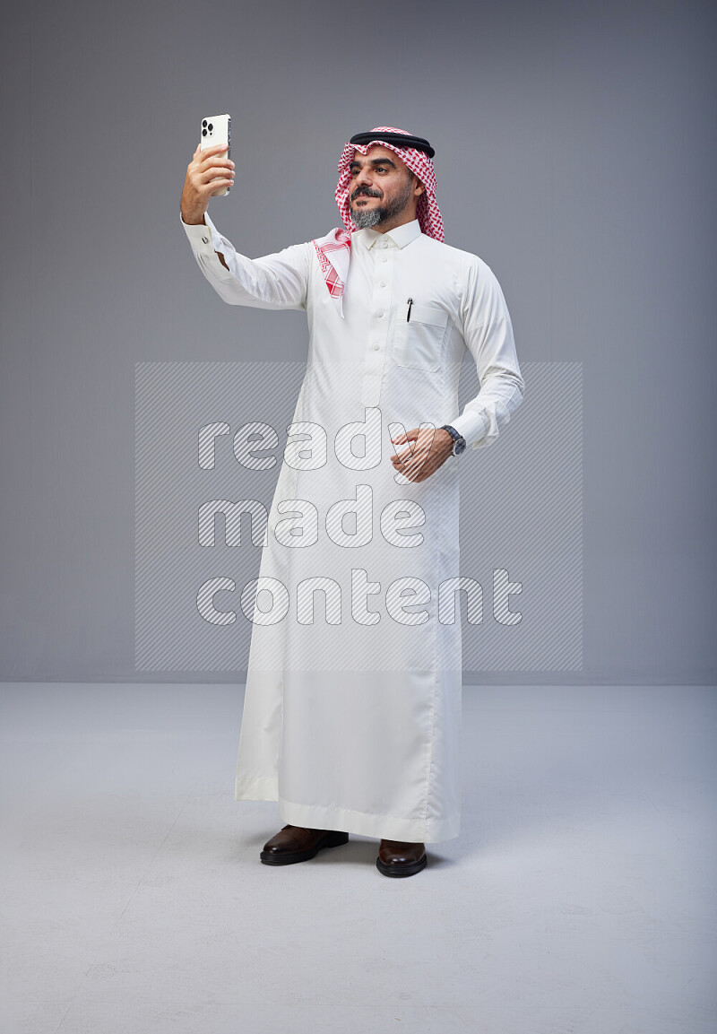 Saudi man Wearing Thob and red Shomag standing taking selfie on Gray background