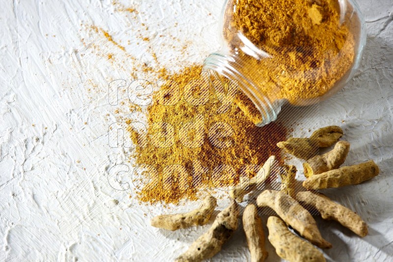 A flipped glass spice jar full of turmeric powder and powder spilled out of it with dried whole fingers on textured white flooring