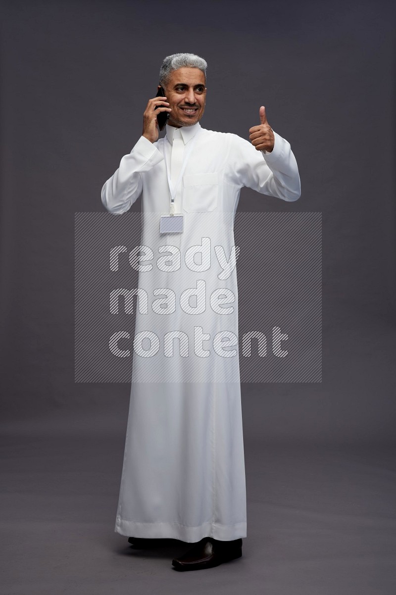 Saudi man wearing thob with neck strap employee badge standing talking on phone on gray background