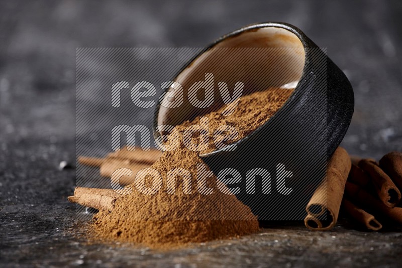 Black pottery bowl over filled with cinnamon powder and cinnamon sticks around the bowl on a textured black background