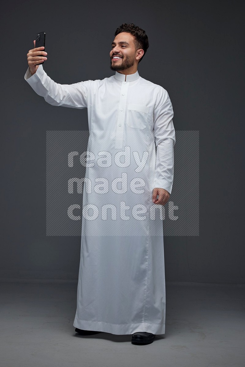 A Saudi man wearing Thobe and taking selfie with his phone making different poses eye level on a gray background