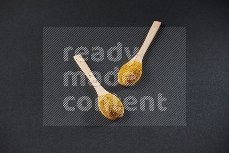 2 wooden spoons full of turmeric powder on black flooring