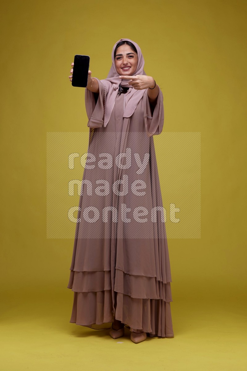 A woman Showing her Phone screen on a Yellow Background wearing Brown Abaya with Hijab