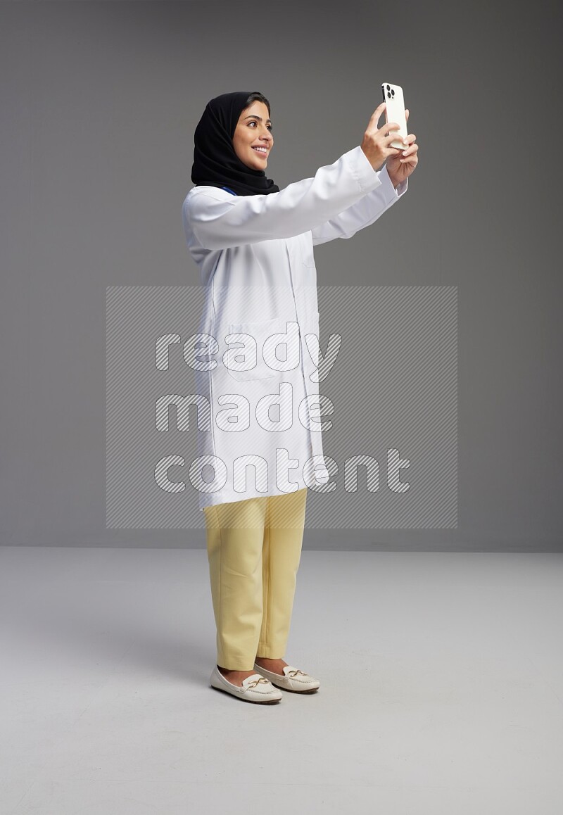 Saudi woman wearing lab coat with stethoscope standing taking selfie on Gray background