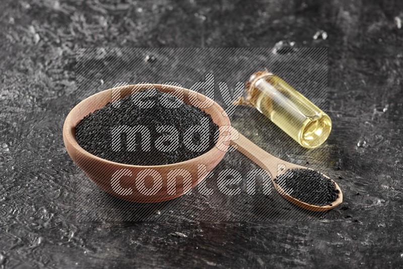 A wooden bowl and spoon full of black seeds and a bottle of black seeds oil on a textured black flooring