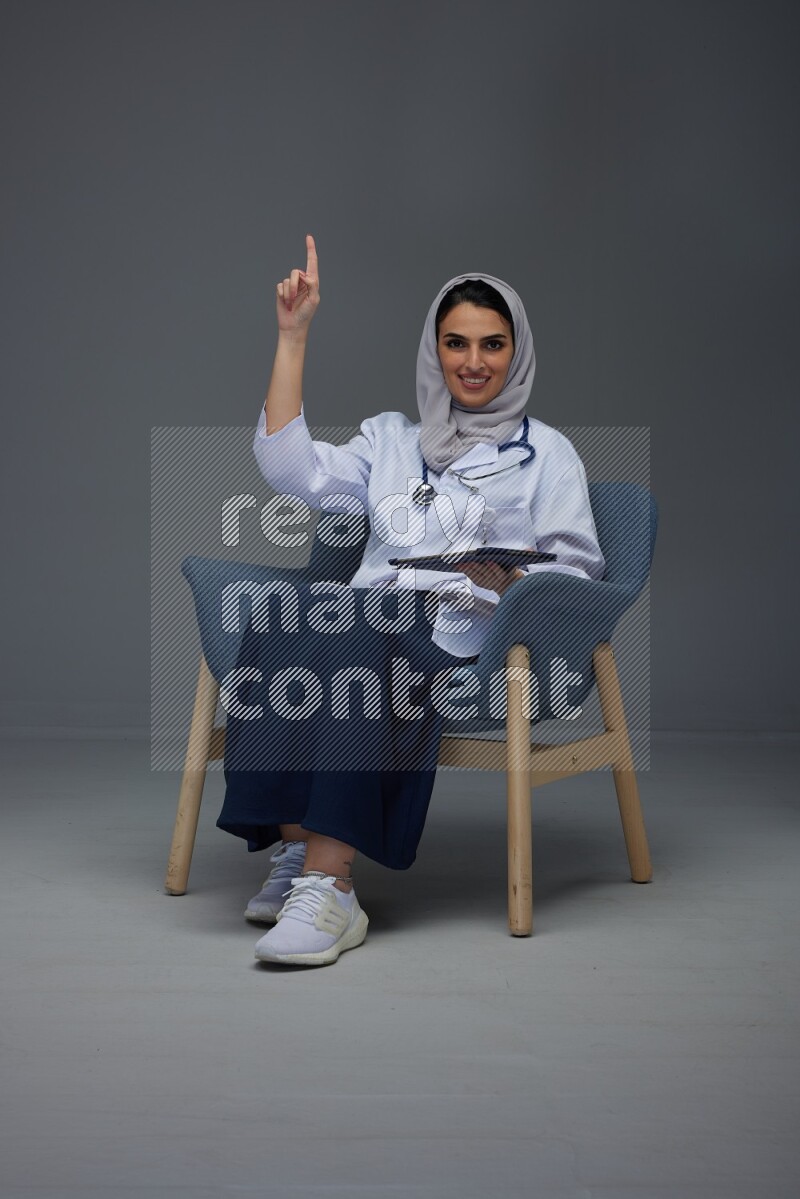 A doctor wearing a light gray head scarf setting on a dark grey chair and using a phone eye level on a grey background