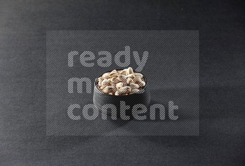 A black pottery bowl full of pistachios on a black background in different angles