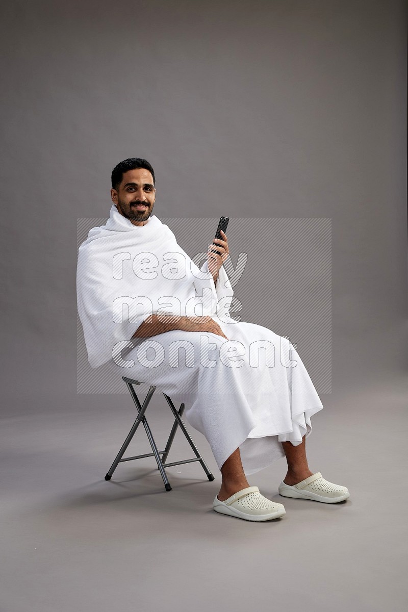 A man wearing Ehram sitting on chair texting on phone on gray background