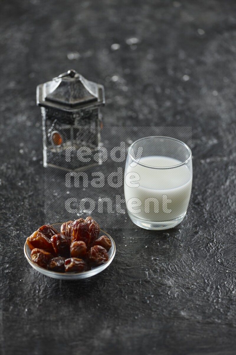 A silver lantern with different drinks, dates, nuts, prayer beads and quran on textured black background