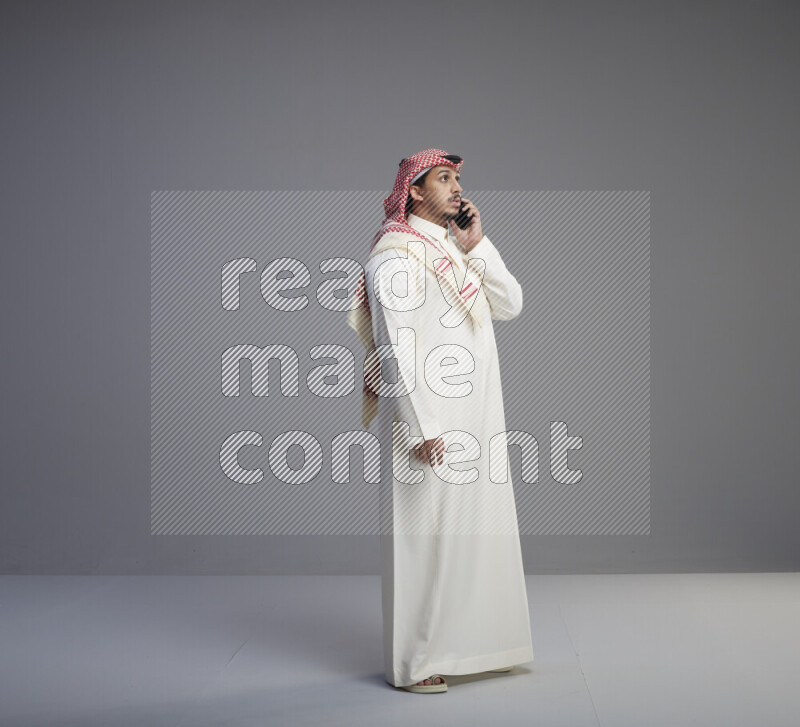 A Saudi man standing wearing thob and red shomag talking on phone on gray background