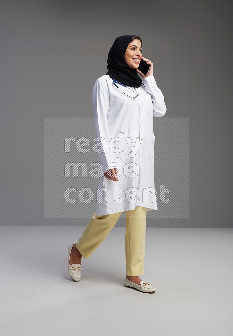 Saudi woman wearing lab coat with stethoscope standing talking on phone on Gray background