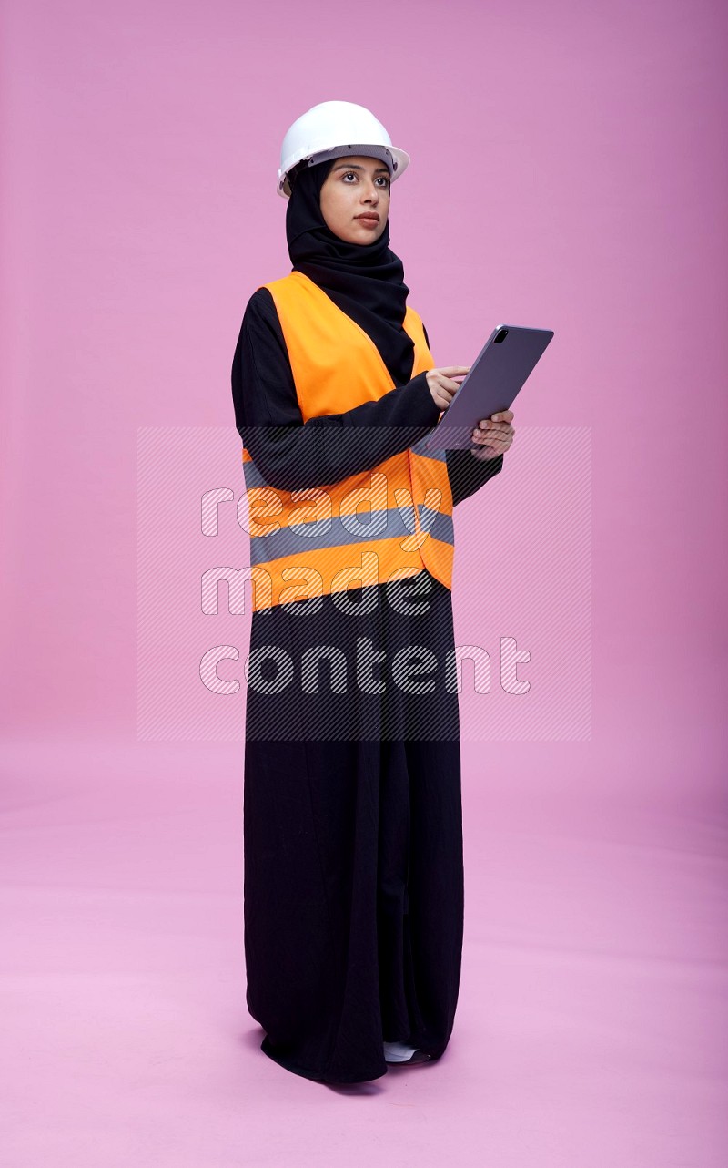 Saudi woman wearing Abaya with engineer vest and helmet standing working on tablet on pink background