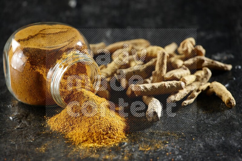 A flipped glass spice jar full of turmeric powder and powder spilled out of it with dried whole fingers on textured black flooring