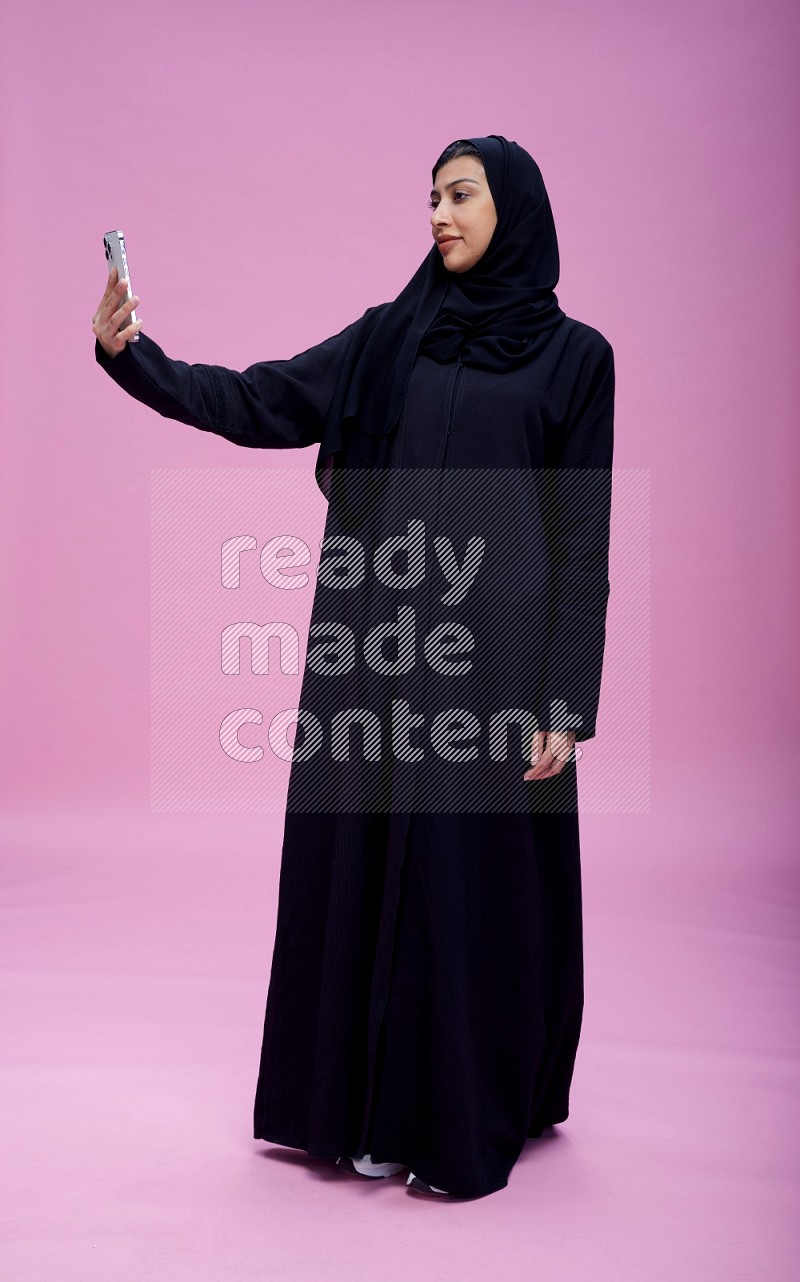 Saudi woman wearing Abaya standing taking selfie on pink background