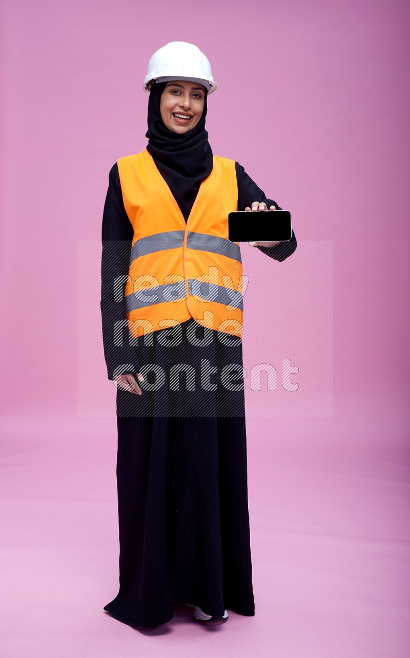 Saudi woman wearing Abaya with engineer vest and helmet standing showing phone to camera on pink background