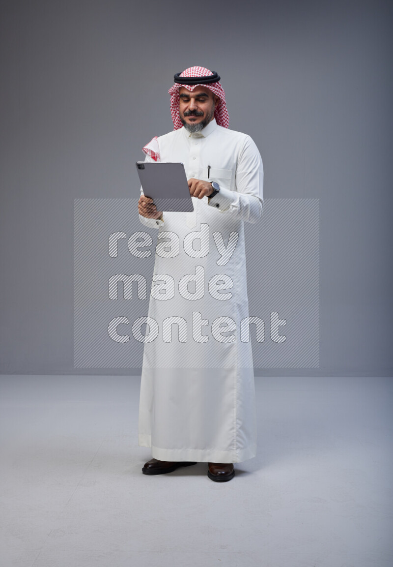 Saudi man Wearing Thob and red Shomag standing working on tablet on Gray background