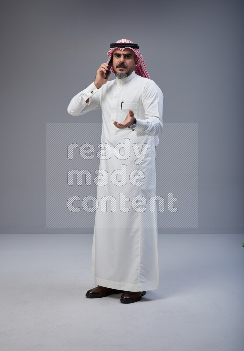 Saudi man Wearing Thob and red Shomag standing talking on phone on Gray background