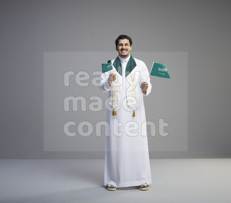 A Saudi man standing wearing thob and saudi flag scarf with face painting holding small Saudi flag on gray background